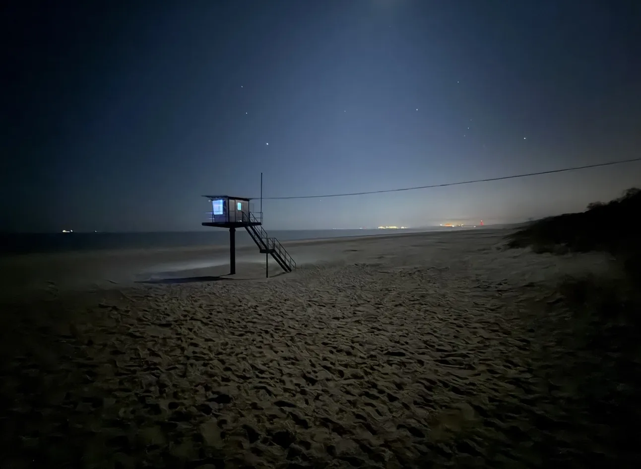 Rettungsschwimmerstation am Ostseestrand bei Nacht: dunkler Sternenhimmel, Sand mit Fußspuren im Vordergrund und Lichter am Horizont