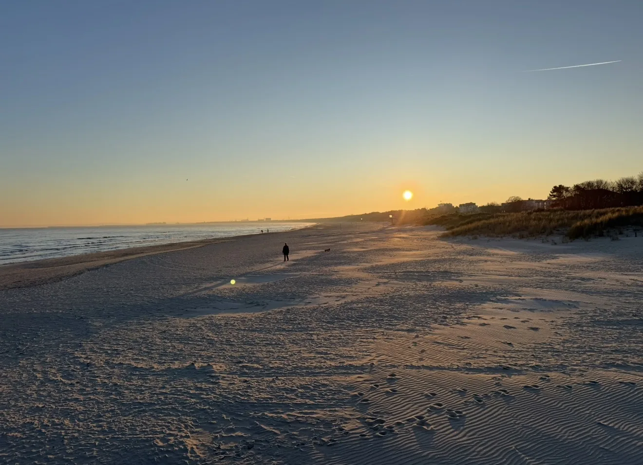 Sonnenuntergang über der Ostsee: die tief stehende Sonne über dem Wasser, Sand mit Wellenrippeln im Vordergrund, Dünen rechts und eine einzelne Person am Strand